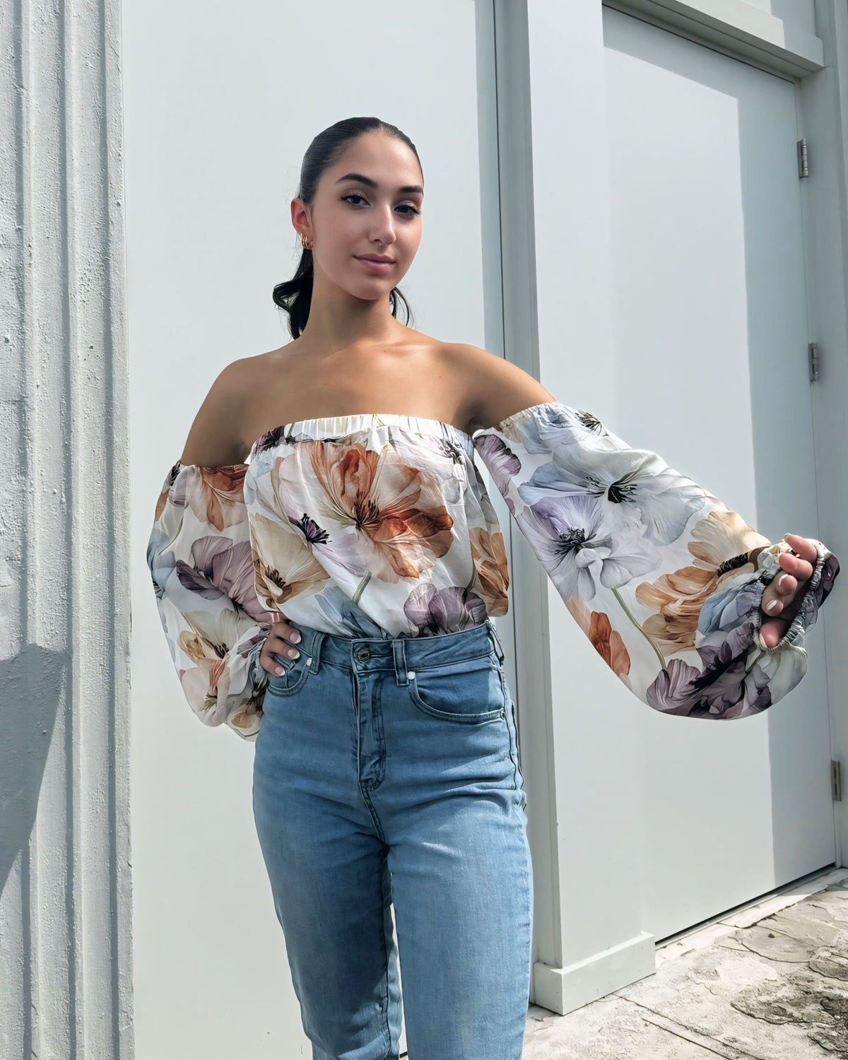 Woman wearing a floral off-shoulder top and jeans standing against a light-colored wall.