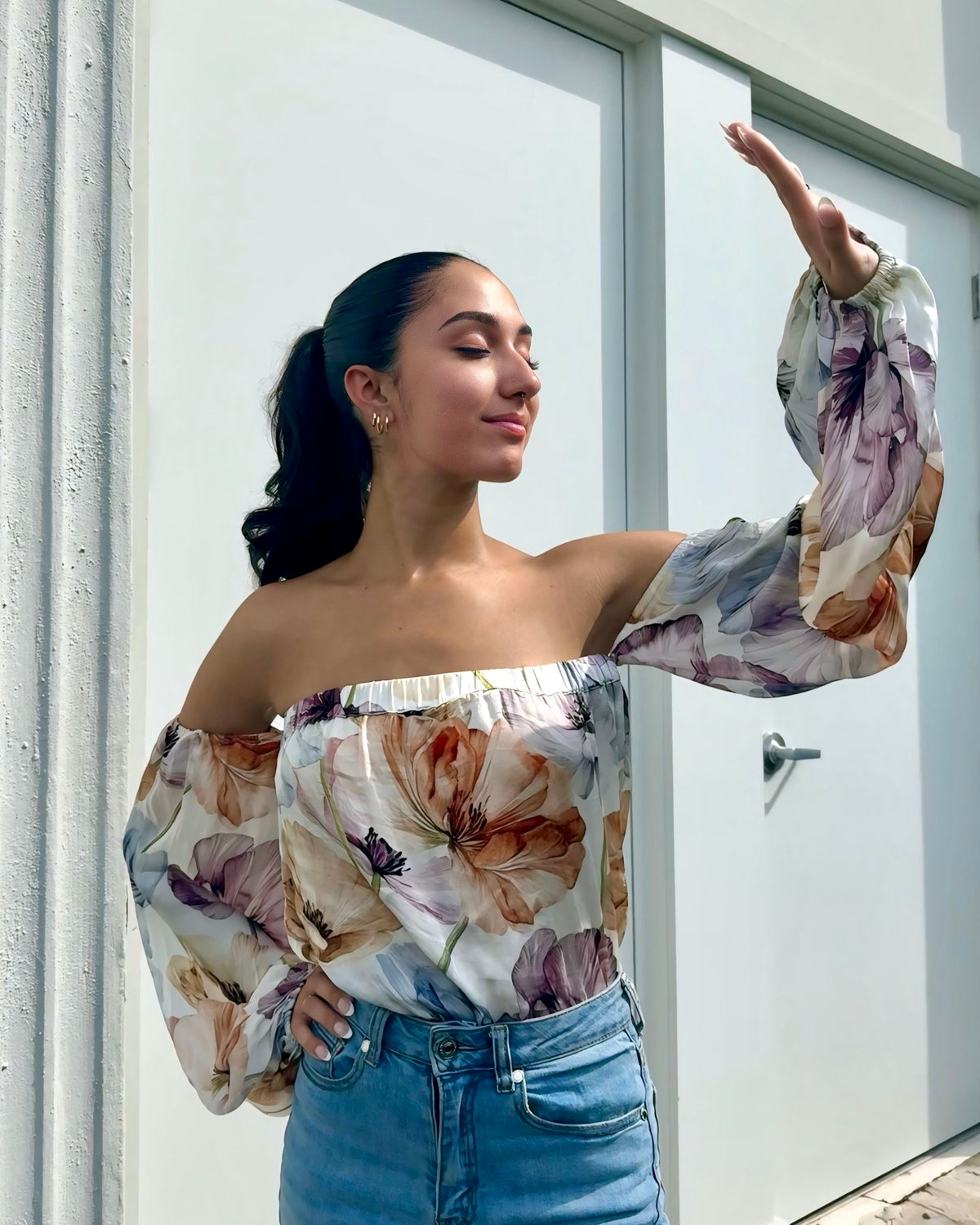 Woman wearing a floral off-shoulder top and blue jeans standing against a white door.
